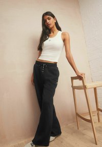 Junge Frau, die ein weißes ärmelloses Oberteil und schwarze weite Sweatpants trägt, lehnt sich in einem minimalistischen Raum an einen Holzstuhl.