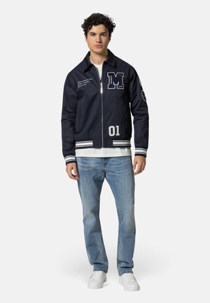 Junger Mann mit lockigem Haar trägt eine marineblaue Varsity-Jacke mit den Aufnähern "M" und "01", hellblaue Jeans, weiße Turnschuhe und darunter ein weißes Hemd.