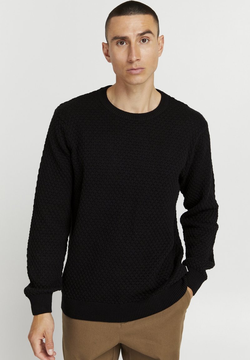 Maglione nero lavorato a maglia con un motivo testurizzato, scollatura tonda e polsini a costine. Abbinato a pantaloni marrone chiaro.