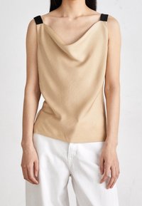 Cowl-neck topp i beige tyg med svarta axelremmar; slät textur med draperad design och en något utflugen fåll. Matchas med vita byxor.