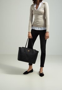 Bolso tote de tejido negro con asas dobles y acento de logotipo dorado, combinado con un suéter beige y leggings negros; zapatos planos negros visibles.
