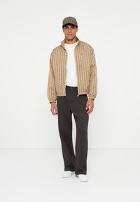 Beige mønstrede bomberjakke med elastiske manchetter, båret over en hvid t-shirt, kombineret med mørke bukser og hvide sneakers, toppet med en ternet kasket.