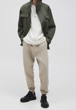 Oliivinvihreä takki, jossa kaksi rintataskua, alla valkoinen pyöreäkauluksinen t-paita, vaaleanbeiget kapealahkeiset collegehousut ja mustat lenkkarit.