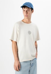 Young man wearing a beige t-shirt with a small sun graphic, light blue jeans, a blue cap, and showing a tattoo on his left forearm.