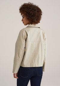 Beige cropped jack met kraag, met een gladde textuur en zichtbare stiksels op de achterkant. Gedragen over donkerblauwe spijkerbroek.
