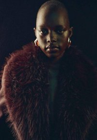 Modella che indossa un elegante scialle scuro simile alla pelliccia di colore borgogna profondo. Orecchini decorativi in oro e rosso molto evidenti. Lo sfondo scuro mette in risalto i tratti.