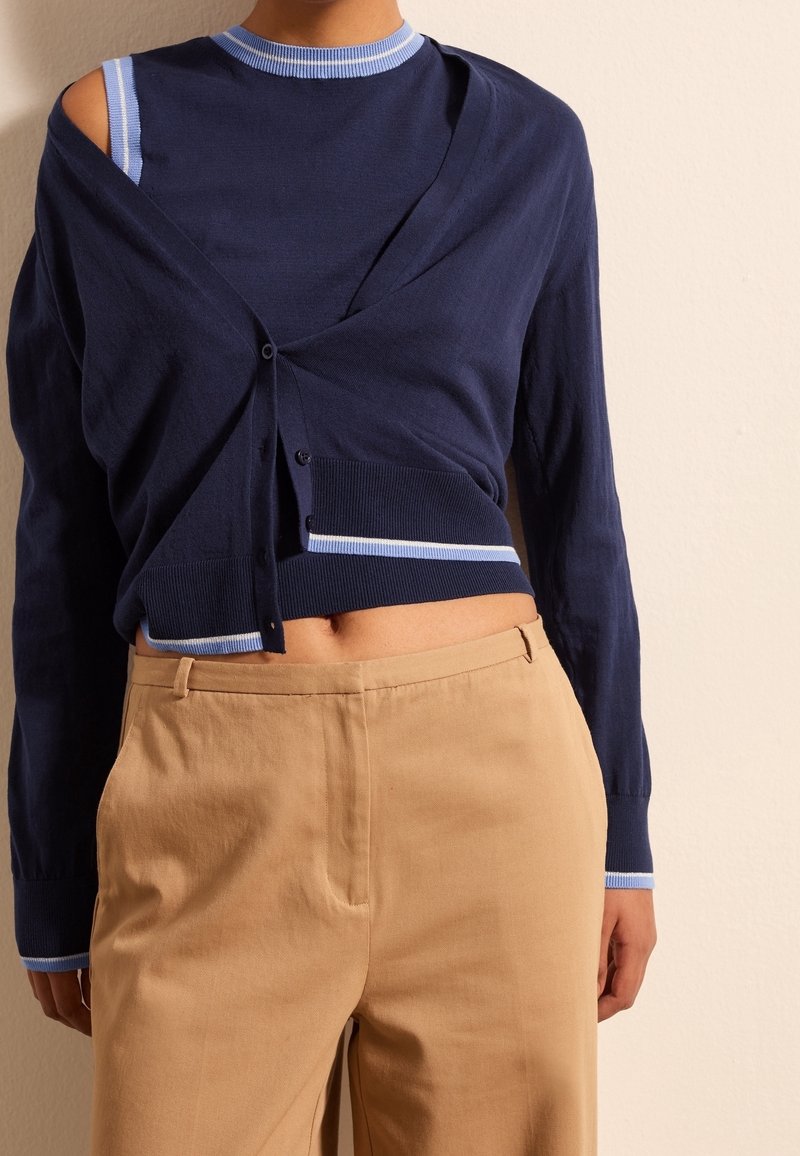 Cardigan blu navy con dettagli azzurri, dotato di scollatura a V, chiusura con bottoni e una spalla scoperta, abbinato a pantaloni beige.