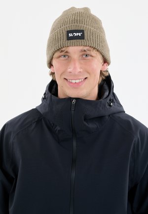 Jong persoon met licht haar die een tan kleurige gebreide beanie draagt met het label "SLOPE" en een zwarte ritsjack met hoge kraag.