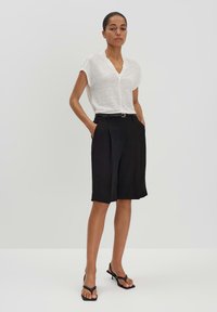 Blusa bianca in lino con scollo a V, abbinata a pantaloni corti neri plissettati e una cintura nera. La modella indossa infradito nere con tacco.