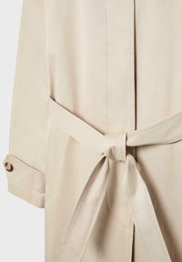 Zone de la taille d'un trench beige avec une ceinture en tissu nouée et un bracelet de manche avec fermeture par bouton.