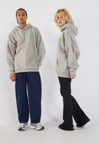 Komfortable grå hettegensere med grønne grafikker båret av to modeller. Den ene i løse denimjeans, den andre i svarte vide bukser.