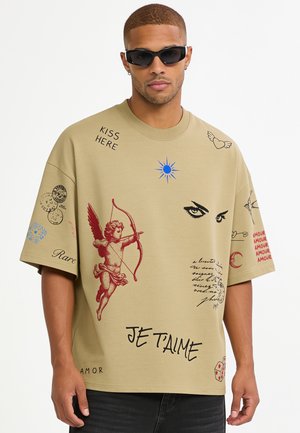 Mann trägt beige T-Shirt mit Amor, Augen, Sonne, Herz mit Flügeln, Würfel und französischem Text einschließlich "Je t'aime" und "Kiss Here," kombiniert mit schwarzen Sonnenbrillen.
