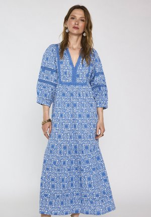 Donna in piedi con un abito lungo a fiori blu e bianchi a maniche lunghe, indossa orecchini e braccialetti su uno sfondo semplice.
