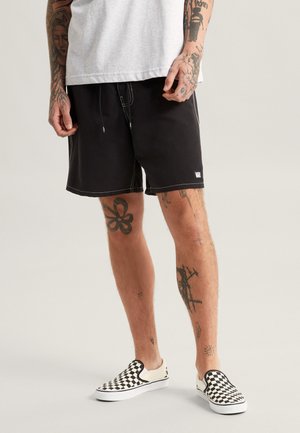 Black swim shorts with white stitching, featuring an elastic waistband with a drawstring. Worn with black and white checkered slip-on shoes.