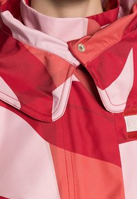 Rød og pink mønstret jakke med høj hals, trykknaplukning og kontrasterende stoftyper med synlige syninger.