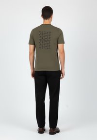 Olivově zelené tričko s krátkým rukávem, s tmavým vzorem a logem na zadní straně; nošeno s černými kalhotami a hnědými botami. Hladká textilní struktura.