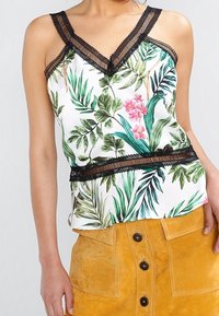 Donna che indossa un top senza maniche bianco con stampa di foglie verdi e fiori rosa, bordo in pizzo nero, e una gonna gialla senape con bottoni frontali.