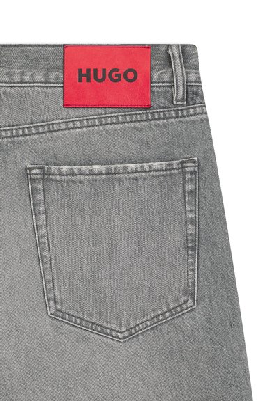 Poche arrière d'un jean en denim gris avec une étiquette rouge cousue au-dessus, affichant le nom de la marque "HUGO" en lettres noires audacieuses.