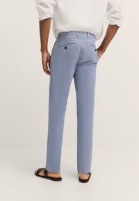 Helle blaue Hose aus glattem Stoff, slim fit mit geradem Schnitt, zwei Gesäßtaschen mit Knopfverschluss, getragen mit schwarzen Sandalen.