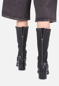 Nohy v zkrácených tmavých džínových kalhotách, obuté v černých kožených botách nad kotníky se zadními zipy, přezkami a nízkými blokovými podpatky na bílém pozadí.