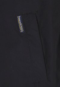 Zwarte stoffen jas met een opgestikt zakje en een blauw label met gele tekst, gestikte randen en een gladde textuur.