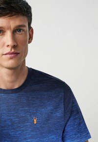 Kék textúrájú póló, kerek nyakkivágással és egy apró narancssárga homár logóval a mellkasán. Rövid ujjai teszik teljessé a dizájnt.