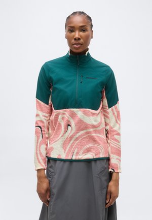 Γυναίκα που φοράει μπλουζάκι με τετράγωνο φερμουάρ σε τιρκουάζ και ροζ σχέδιο και γκρι φούστα, στέκεται μπροστά σε απλό λευκό φόντο.