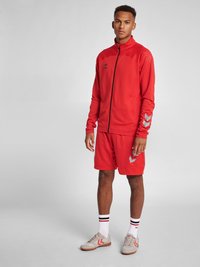Rød atletisk træningsdragt med en jakke med lynlås og shorts. Jakken har en høj krave og prikket mønster; shortsene har et kontrasterende logo.