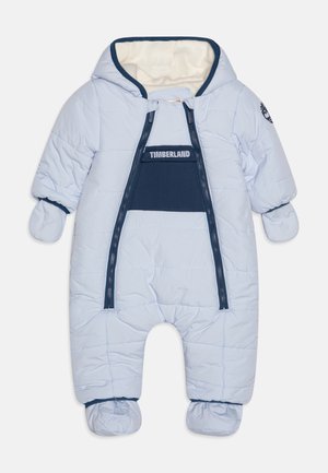 Tuta da neve imbottita azzurro chiaro con cappuccio foderato in pile, chiusura con zip frontale e polsini elastici. Presenta un pannello di dettaglio blu navale e maniche coperte da guanti.