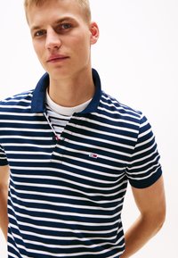 Marinblå och vit randig polo t-shirt med krage, prydd med en liten broderad logotyp på vänster sida. Korta ärmar, mjuk textur.