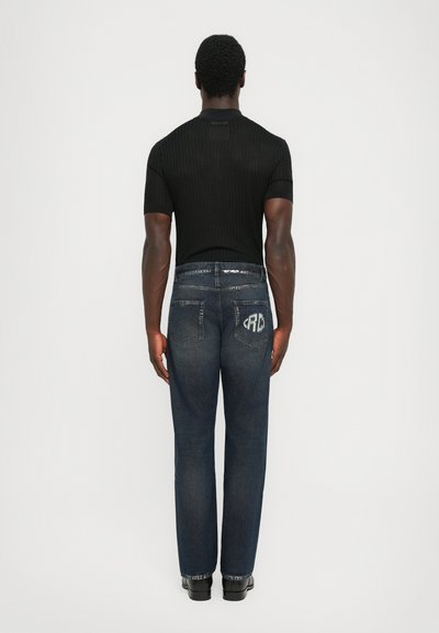 Man standing with back to camera wearing a black short-sleeve shirt and dark blue jeans with a white logo on the right back pocket.