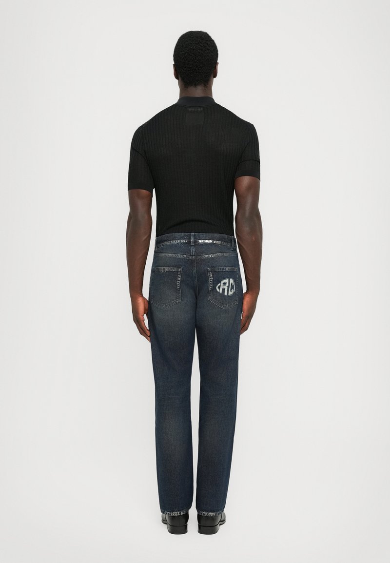 Uomo in piedi con le spalle alla fotocamera, indossa una maglietta nera a maniche corte e jeans blu scuro con un logo bianco sulla tasca posteriore destra.