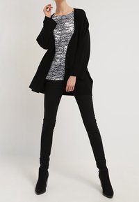 Woman wearing black buttoned cardigan, black pants, black ankle boots, and a patterned black-and-white top, standing with legs apart.