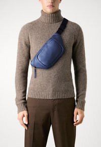Μπλε δερμάτινη τσάντα σέλας φορεμένη πάνω από ένα καφέ πουλόβερ με ψηλό λαιμό από μάλλινο υλικό. Η τσάντα έχει ορθογώνιο σχήμα, κλείσιμο με φερμουάρ και ανάγλυφο λογότυπο.