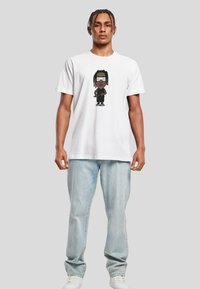 White cotton t-shirt featuring a graphic design of a cartoon character in black attire and sunglasses; model paired with light blue jeans.