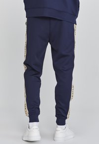 Marineblauwe joggingbroek heeft een taps toelopend ontwerp met contrasterende beige zijstrepen waarop de merknaam in goud is weergegeven. Draag ze met witte sneakers.