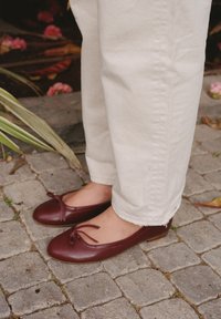 Burgundy nahkaa ballet-kengät pyöreällä varpaalla ja nauhadetalla, yhdistettynä kerma värisiin, käännettyihin housuihin kivisellä polulla.