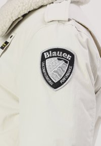 Balts žakets ar vilnas apkakli, ar melnu un sudraba iešūtu uzpleca uzšuvumu, kas marķēts ar "Blauer". Gluda tekstūra.