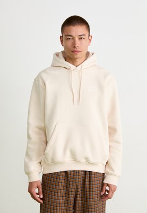 Cream-colored hoodie made of soft fabric, featuring a front kangaroo pocket and a small logo on the chest. Worn with checked pants.