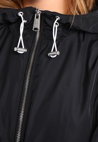 Czarna kurtka z zamkiem błyskawicznym i kapturem, z elastycznymi sznurkami regulacyjnymi, błyszczący srebrny zamek oraz gładki, lekki materiał.