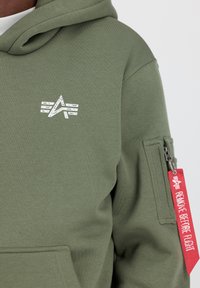 Hanorac verde olivescu cu logo alb pe piept, material texturat și buzunar cu fermoar pe mânecă, echipat cu un eticheta roșie "REMOVE BEFORE FLIGHT".