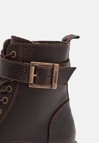 Bottines en cuir marron avec une finition texturée, une sangle de boucle sur le côté, des œillets en métal, et un tirant contrastant rouge et noir.