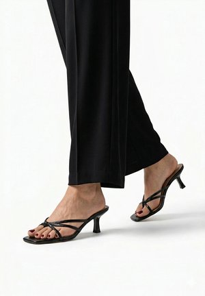 Pies con sandalias negras de tacón alto y tiras, con pantalones negros, mostrados sobre un fondo claro y liso.