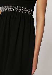 Robe noire avec une ceinture ornée de pierres précieuses transparentes de formes variées, portée sur le bras et le torse d'une personne sur un fond clair.
