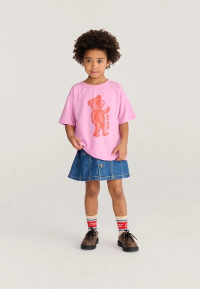 Enfant aux cheveux bouclés portant un t-shirt rose avec un motif ours orange, une jupe en jean, des chaussettes colorées et des chaussures marron à lacets, debout sur un fond clair.