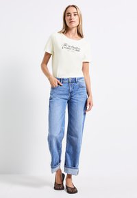 T-shirt en coton jaune clair avec texte noir, associé à un jean en denim bleu clair, ample, avec les manches retroussées et des ballerines en mesh noires.