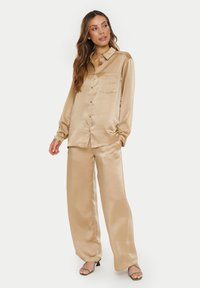 Beige satin shirt with a collared neckline, front pocket, button closure; paired with wide-leg satin trousers. Green heeled sandals complete the look.