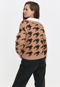 Beżowy puszysty sweter w czarno-białą kratkę houndstooth, z okrągłym dekoltem i opadającymi ramionami. Noszony na ciemnych dżinsach.