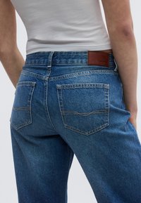 Dark blue denim jeans with a high waistband, featuring two back pockets and a brown leather patch on the waist with logo detail.