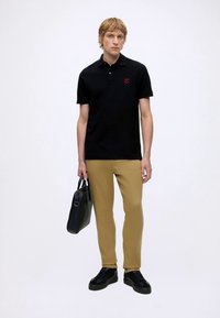 Young man wearing a black polo shirt, tan pants, black sneakers, holding a black briefcase against a plain white background.